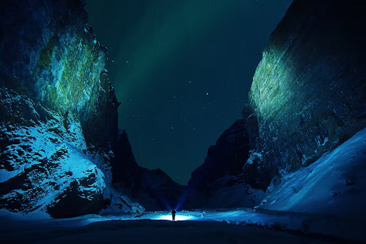 A hiker stands at the bottom of snowy Stakkholtsgja Canyon, Iceland at night with flashlights. 