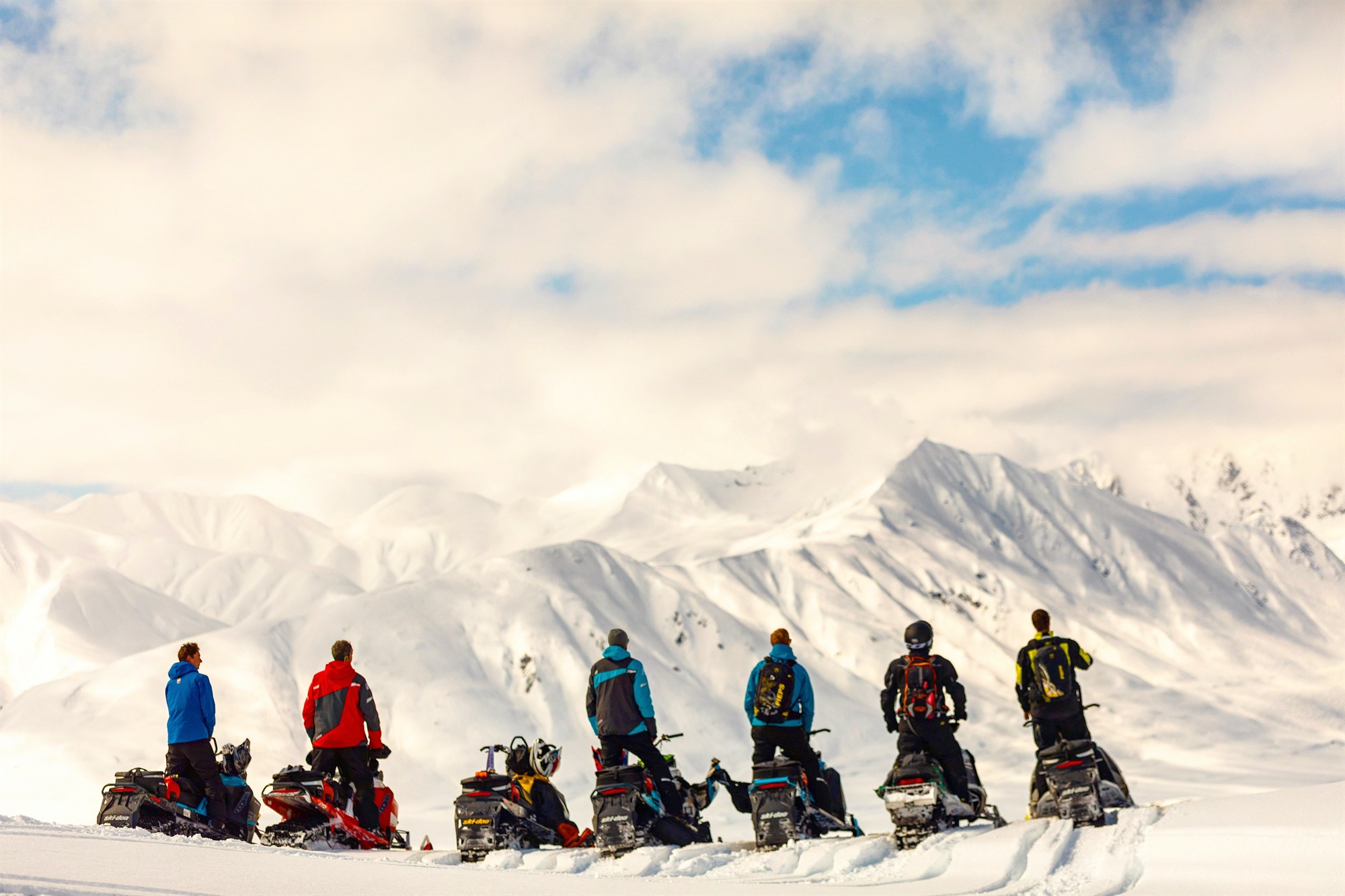 Row of people on snowmobiles looking at a snowy mountain range.